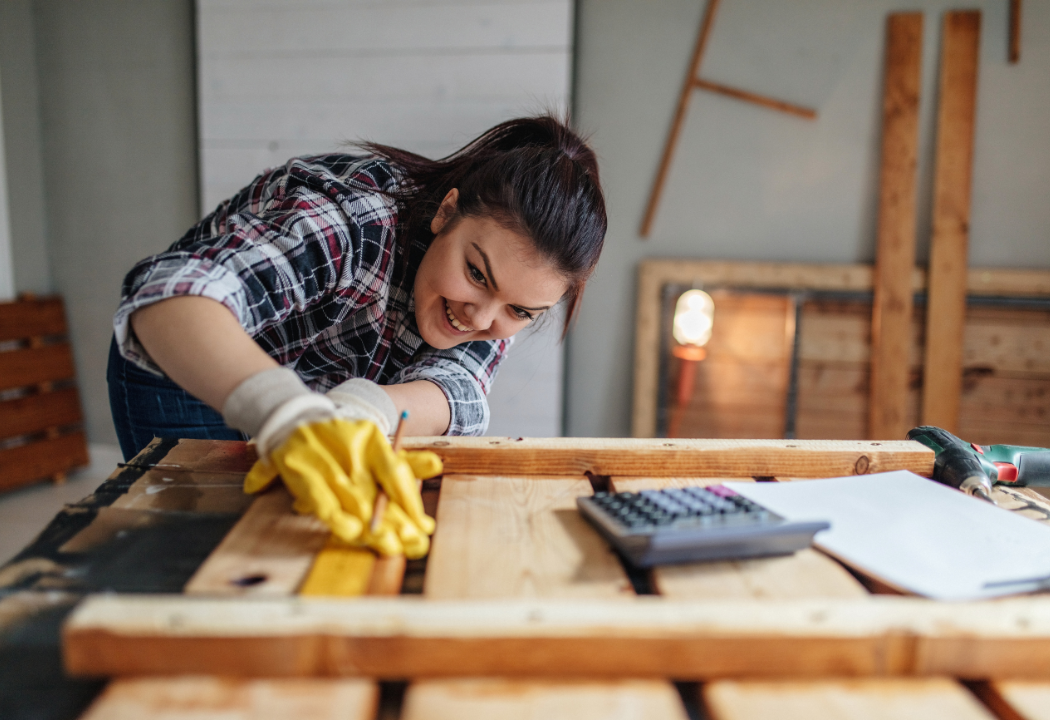 A woman working on a work bench measuring a piece of wood. 